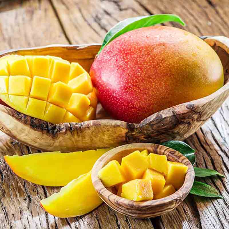 Eine frische Mango und gewürfelte Stücke liegen in einer Holzschale auf einer rustikalen Oberfläche, akzentuiert durch leuchtend grüne Blätter. Eine kleine Schale, ideal für Cocktails, präsentiert weitere gewürfelte Mango. Diese exotische Präsentation ist perfekt für alle, die tropische Aromen mit Mango Balsamessig von Acetaia Murani suchen.