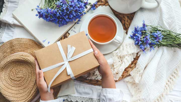 Geschenkbox, Tee, Blumen, Strohhut, weiße Tischdecke, gemütliche Atmosphäre.